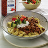 A bowl of pasta with ground meat tomatoes cucumbers and a dollop of sour cream with a McCormick Culinary Crushed Red Pepper container in the background.