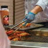 A person in a white chef coat uses tongs to flip a lobster tail on a grill next to a bottle of McCormick Grill Mates Seafood Seasoning.