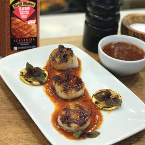 A plate of grilled scallops with lemon and basil topped with a red sauce and a small bowl of sauce in the background.