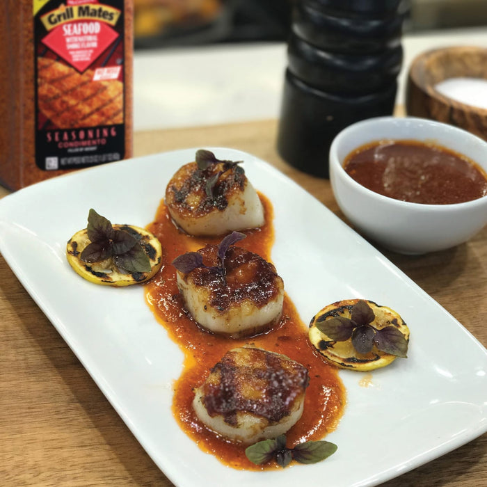 A plate of grilled scallops with lemon and basil topped with a red sauce and a small bowl of sauce in the background.