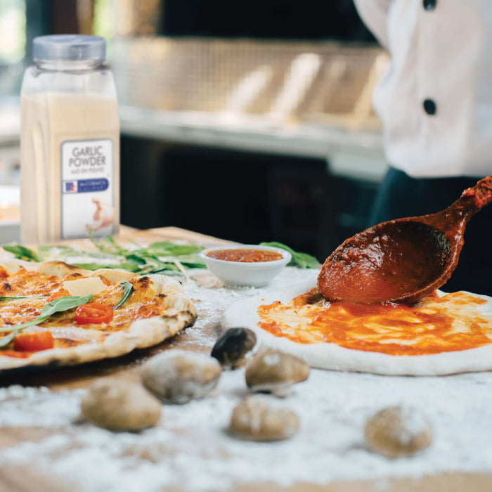 A wooden spoon is spreading tomato sauce on a pizza crust with a finished pizza and a jar of McCormick Garlic Powder in the background.