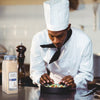 A chef in a white uniform is plating a dish of food with a jar of McCormick Culinary Granulated Garlic in the foreground.