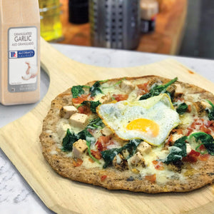 A close-up of a pizza with spinach chicken bacon and a fried egg on a wooden cutting board next to a McCormick Culinary Granulated Garlic container.