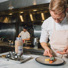 A chef in a white coat and apron is plating a dish of lobster with a side of corn puree and greens.