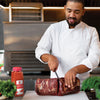 A chef in a white coat is cutting a large piece of meat with a knife next to a jar of Lawrys Memphis Style Barbecue Rub.