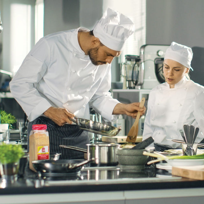 Two chefs in white uniforms are cooking in a kitchen one is holding a pan and the other is holding a wooden spoon.