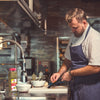 A chef in a white shirt and blue apron is preparing food in a kitchen with a Lawrys All Purpose Recipe Blend Seasoning jar on the counter.