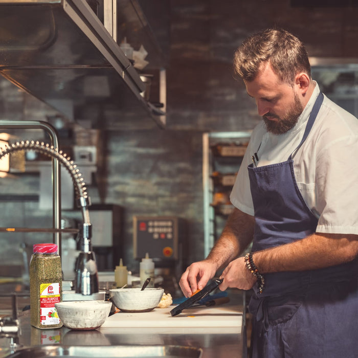 A chef in a white shirt and blue apron is preparing food in a kitchen with a Lawrys All Purpose Recipe Blend Seasoning jar on the counter.