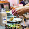 A hand holds chopsticks over a plate of food with a McCormick Culinary Whole Sesame Seed container in the foreground.