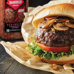 A hamburger with mushrooms and onions on a bun sitting on brown paper next to a jar of Hamburger Seasoning.