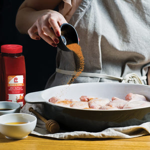 A hand sprinkles Lawrys Spicy Buffalo Wings Seasoning Mix over raw chicken in a bowl.