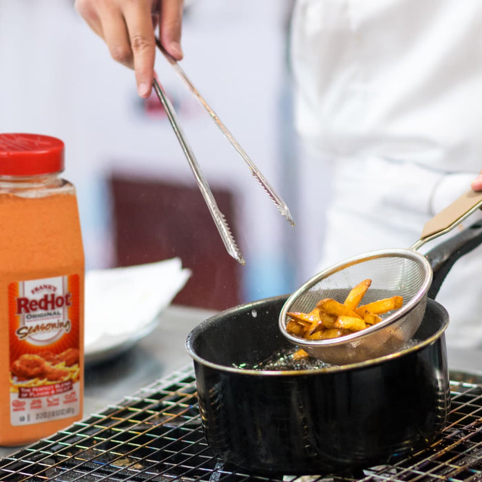 A person is using tongs to remove french fries from a pot of hot oil with a bottle of RedHot Original Seasoning in the foreground.