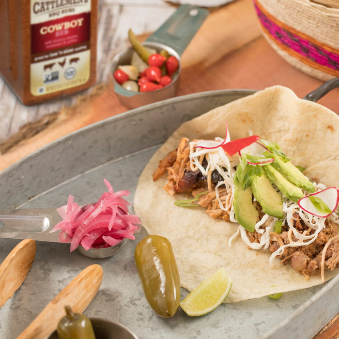 A close-up of a pulled pork taco with avocado radish and pickled jalapeno with Cattlemens BBQ Rub in the background.