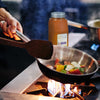 A hand holds a spatula over a pan of vegetables cooking on a stovetop with a jar of Moroccan seasoning in the background.