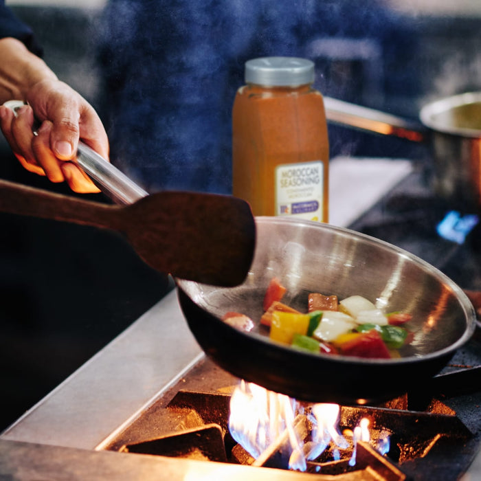 A hand holds a spatula over a pan of vegetables cooking on a stovetop with a jar of Moroccan seasoning in the background.