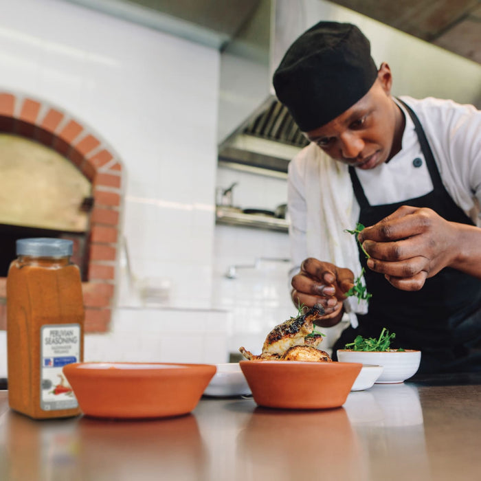 A chef in a white chefs coat and black apron is garnishing a plate of food with fresh herbs.