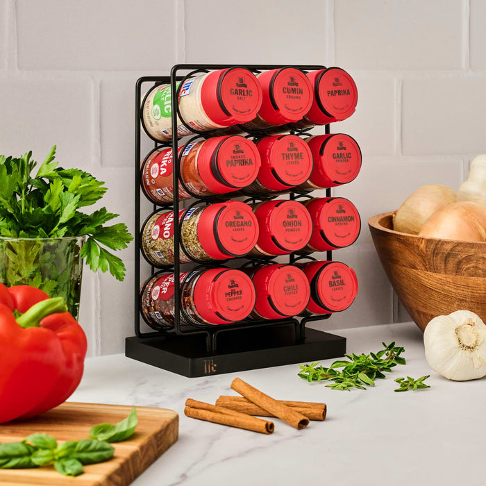 A Kamenstein spice rack with filled spice jars sits on a kitchen counter next to fresh produce.