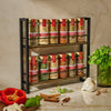 A two-tiered spice rack filled with labeled spice jars sits on a kitchen counter alongside fresh ingredients like peppers and herbs.