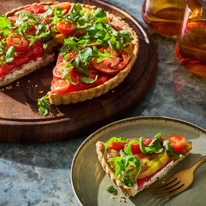 A vibrant tomato tart topped with fresh greens sits on a wooden board, with a slice plated nearby, suggesting a delicious and visually appealing dish.
