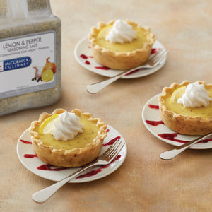 Three lemon tarts with whipped cream on top a fork and a jar of McCormick Culinary Lemon &amp; Pepper Seasoning Salt.