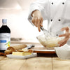 A chef is mixing ingredients in a bowl with a whisk with a bottle of McCormick Culinary Pure Vanilla Extract a rolling pin a knife and a dish of butter on a wooden table.