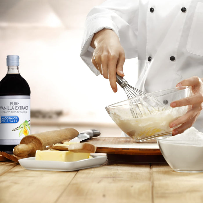 A chef is mixing ingredients in a bowl with a whisk with a bottle of McCormick Culinary Pure Vanilla Extract a rolling pin a knife and a dish of butter on a wooden table.