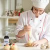 A chef in a white uniform with red trim is decorating a cupcake with a piping bag while a bottle of McCormick Culinary Imitation Vanilla Flavor sits on the table.