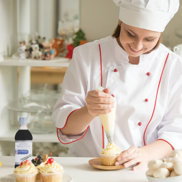 A chef in a white uniform with red trim is decorating a cupcake with a piping bag while a bottle of McCormick Culinary Imitation Vanilla Flavor sits on the table.