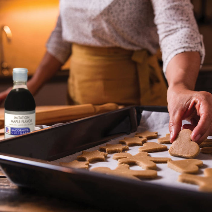 A person is placing a heart-shaped cookie on a baking sheet with other cookies next to a bottle of McCormick Culinary Imitation Maple Flavor.