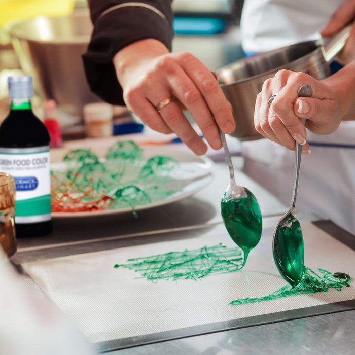 A person is using a spoon to drizzle green food coloring onto a baking sheet.