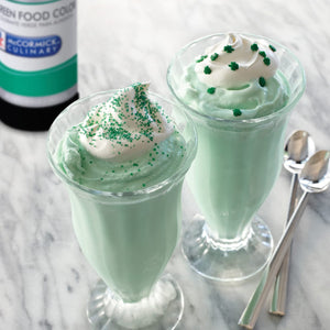 Two glasses of green mint milkshake topped with whipped cream and green sprinkles with two silver spoons on the side.
