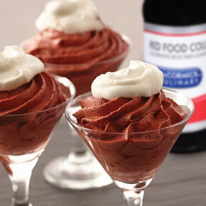 Three glasses of chocolate mousse topped with whipped cream are on a table next to a bottle of McCormick Red Food Color.