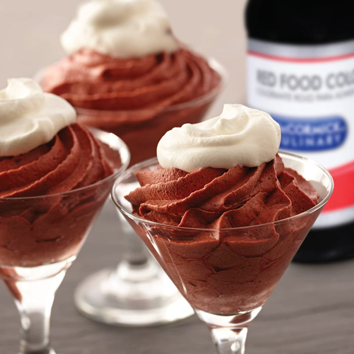 Three glasses of chocolate mousse topped with whipped cream are on a table next to a bottle of McCormick Red Food Color.
