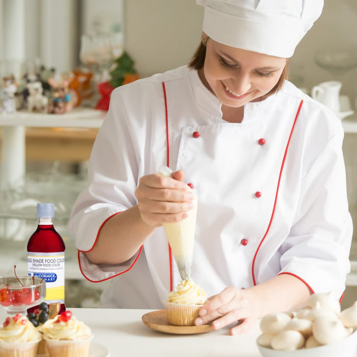 A baker in a white chefs coat and hat decorates a cupcake with frosting while holding a piping bag in one hand and a bottle of McCormick Egg Shade Food Color in the other.