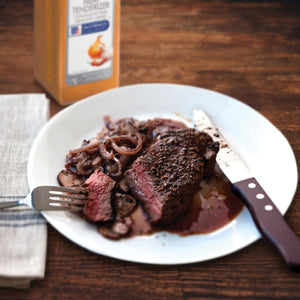 A plate of a steak with mushrooms and onions a fork a knife and a bottle of McCormick Tenderizer.