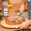 A person is holding a wooden cutting board with a pumpkin pie on it and a jar of McCormick Pumpkin Pie Spice is in the background.