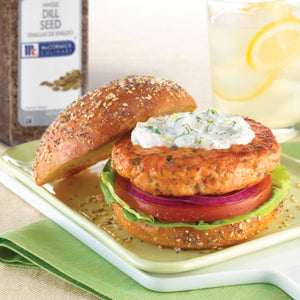 A McCormick Dill Seed spice jar is next to a salmon burger on a sesame seed bun with a creamy dill sauce tomato red onion and lettuce.