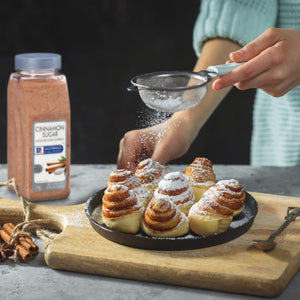 A hand sprinkles powdered sugar over a plate of cinnamon rolls with a bottle of McCormick Cinnamon Sugar in the background.