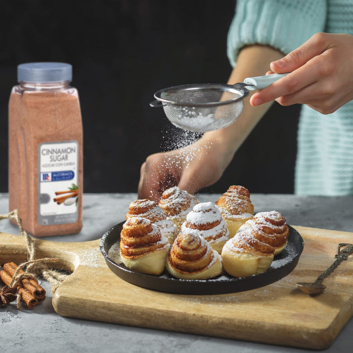 A hand sprinkles powdered sugar over a plate of cinnamon rolls with a bottle of McCormick Cinnamon Sugar in the background.
