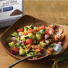 A wooden bowl filled with a fresh cucumber salad with tomatoes red onion and cilantro with a McCORMICK CULINARY BLACK PEPPER container in the background.