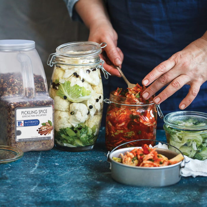 A persons hand reaches into a jar of McCormick Culinary Pickling Spice next to jars of pickled cauliflower and kimchi.
