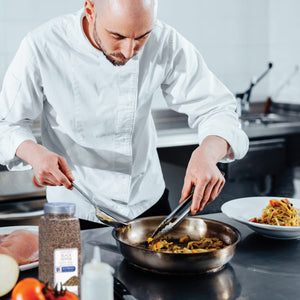 A chef in a white uniform uses tongs to stir vegetables in a pan.