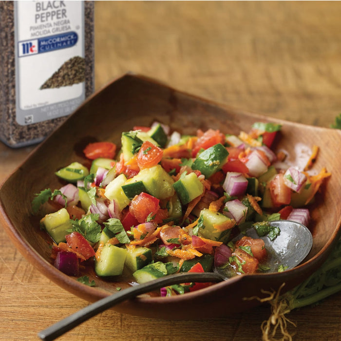 A wooden bowl filled with a fresh cucumber and tomato salad with a spoon and a McCormick Culinary Black Pepper container.