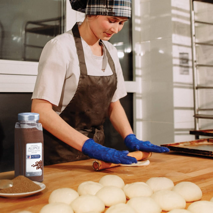 A baker wearing blue gloves rolls out dough on a wooden counter with a jar of McCormick Ground Allspice in the background.