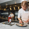 A chef in a white coat and apron is plating a dish of lobster with a side of corn and greens.