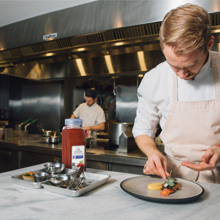 A chef in a white coat and apron is plating a dish of lobster with a side of corn and greens.