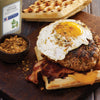 A waffle topped with a sausage patty bacon a fried egg and cheese with a McCormick Ground Thyme bottle in the background.