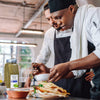 A chef is adding a drizzle of olive oil to a plate of food with a jar of McCormick Culinary Oregano Leaves in the background.