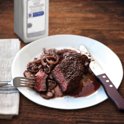 A plate of a juicy steak with caramelized onions and mushrooms a fork a knife and a bottle of McCormick Meat Tenderizer.