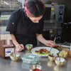 A chef is plating a dish of octopus with green sauce and garnishes using a pair of tongs to arrange the octopus.